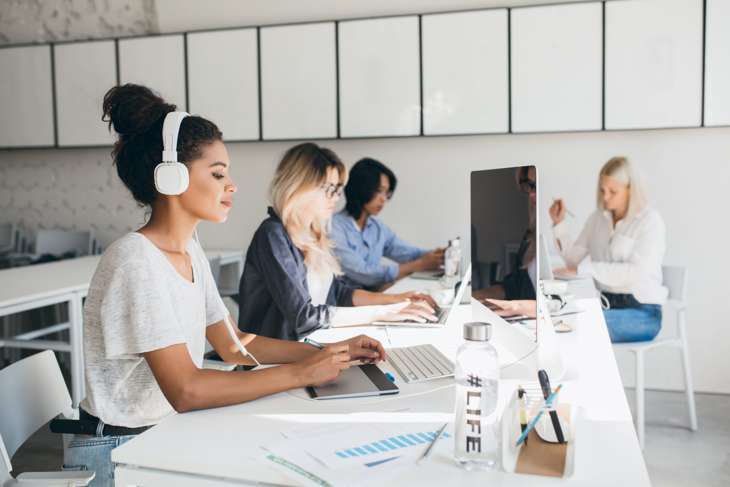 concentrated african female web designer using graphics tablet while her colleagues writing reports. indoor portrait of programmers of international company spending time together at workplace..